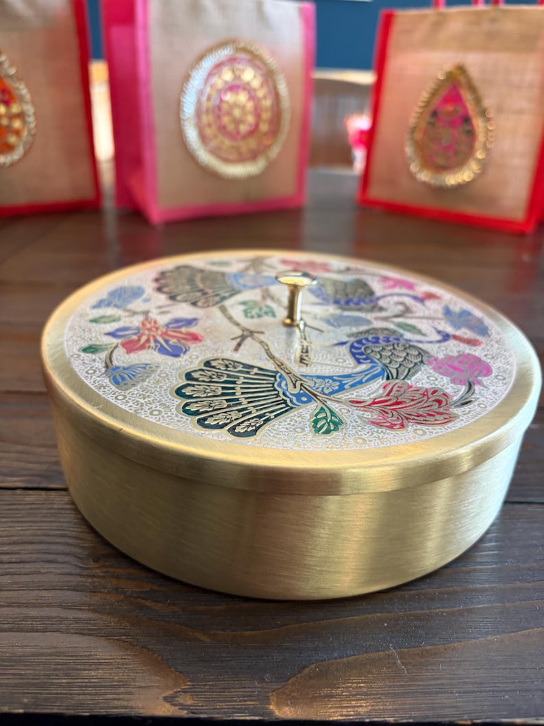 May include: A round, brass-colored container with a decorative lid featuring a peacock and floral design in blue, red, and green. The container sits on a dark wooden surface. In the background are pink and tan gift bags.