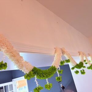 May include: A decorative garland with white and green floral elements. The garland is draped across a doorway, with white, textured sections and vibrant green accents. The design includes hanging green floral clusters.