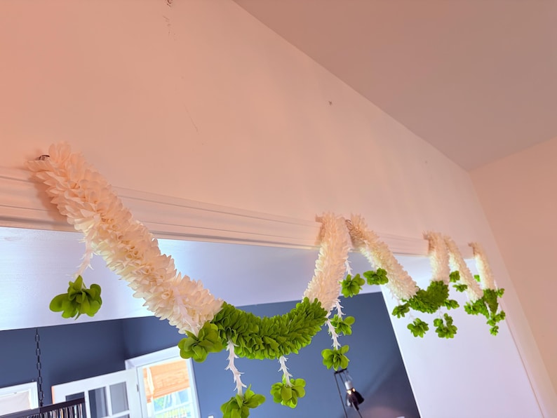 May include: A decorative garland with white and green floral elements. The garland is draped across a doorway, with white, textured sections and vibrant green accents. The design includes hanging green floral clusters.
