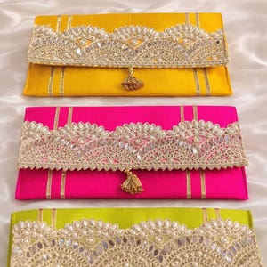 May include: Three rectangular envelope clutches in bright yellow, hot pink, and lime green satin fabric. Each clutch features a wide, scalloped flap adorned with intricate silver embroidery, small mirrors, and a central dangling metallic tassel.