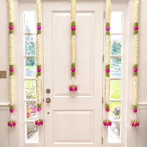 May include: White door decorated with floral garlands. Each garland features white flowers, green accents, and pink spherical ornaments. The garlands hang vertically, framing the door and windows, with a decorative floral cluster at the bottom.