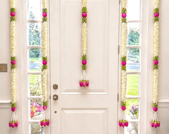 Puede incluir: Puerta blanca decorada con guirnaldas florales. Cada guirnalda presenta flores blancas, detalles verdes y adornos esféricos rosas. Las guirnaldas cuelgan verticalmente, enmarcando la puerta y las ventanas, con un racimo floral en la base.