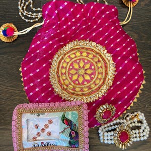 May include: A magenta drawstring pouch with a white diagonal stripe pattern, featuring a large gold circular medallion with mirror accents. Included are a rectangular package of bindi stickers with a peacock illustration and a pearl bracelet.