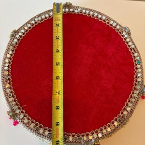 K&ouml;nnte beinhalten: Ein rundes Tablett aus rotem Samt mit einem goldfarbenen Zierrand, der mit kleinen eingelassenen Spiegeln versehen ist. Ein gelbes Stanley-Ma&szlig;band liegt in der Mitte und zeigt einen Durchmesser von etwa 26,7 cm f&uuml;r die Tablettfl&auml;che an.
