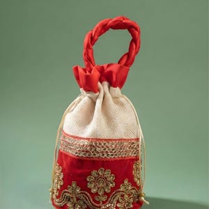 May include: A drawstring pouch bag featuring a cream-colored textured upper section and a vibrant red velvet lower section. The bag is adorned with gold metallic embroidery and sequins in floral patterns, finished with a braided red fabric handle.