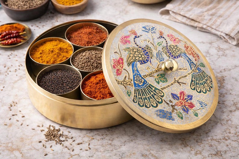 May include: A gold-colored spice box with a decorative lid featuring two peacocks and floral designs. The box contains several compartments filled with various spices, including turmeric, cumin, and chili powder.