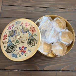 May include: A gold-colored, round decorative box with a lid featuring a peacock design in blue, green, red, and gold. The box is open, revealing several small, gold-colored containers and spoons.
