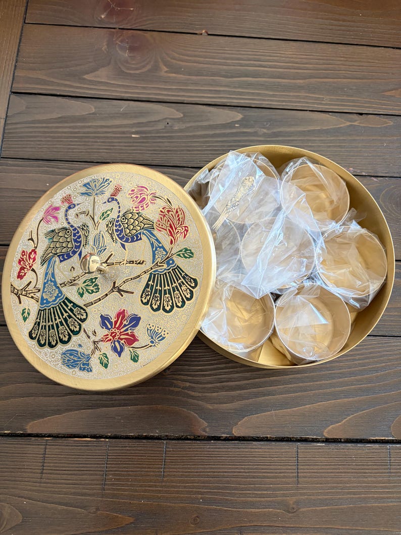 May include: A gold-colored, round decorative box with a lid featuring a peacock design in blue, green, red, and gold. The box is open, revealing several small, gold-colored containers and spoons.