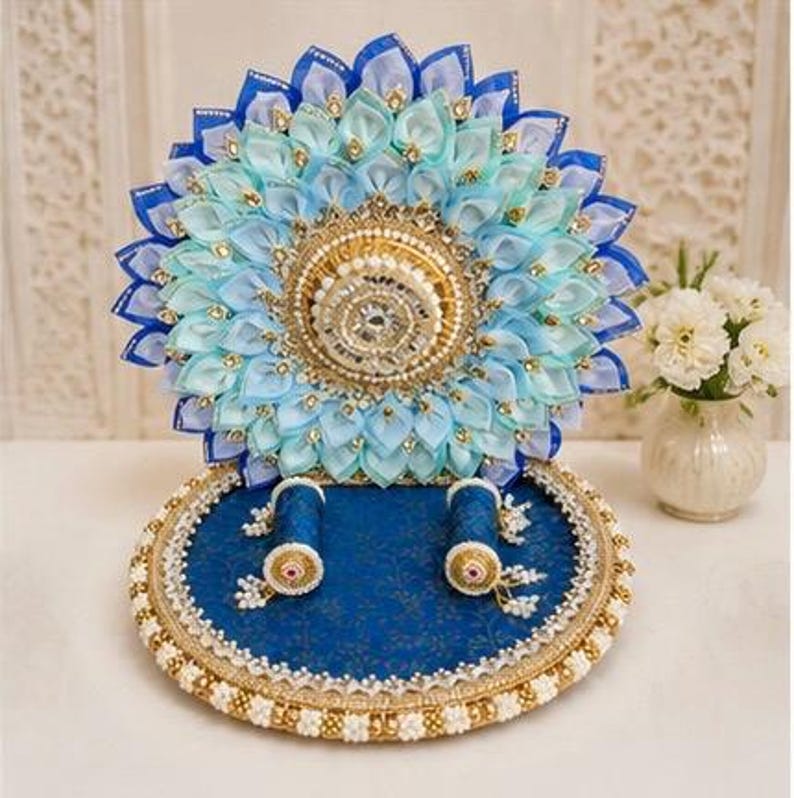Puede incluir: Una bandeja circular azul para exhibir anillos con un panel trasero decorativo hecho de p&eacute;talos de tela en azul claro y azul real. La base incluye dos soportes cil&iacute;ndricos para anillos y est&aacute; adornada con bordes dorados y cuentas blancas.