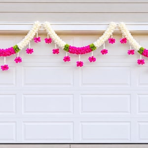 May include: White garage door decorated with three floral garlands. Each garland features white, pink, and green flowers. The garlands are draped across the top of the garage door, creating a festive and decorative display.
