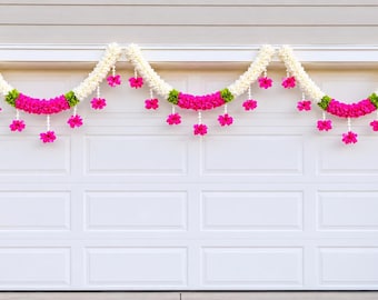 May include: White garage door decorated with three floral garlands. Each garland features white, pink, and green flowers. The garlands are draped across the top of the garage door, creating a festive and decorative display.