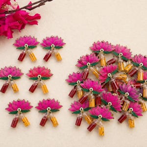 May include: A collection of lotus flower ornaments, each featuring a vibrant pink and green layered design. Attached to the base of each lotus are two miniature glass vials, one filled with red powder and the other with yellow powder.