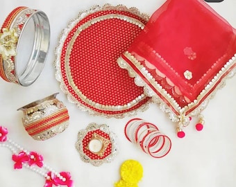 May include: A red and gold ceremonial set including a circular sieve, a decorative plate, a sheer red veil with gold trim, a small metal pot, a candle holder, a stack of red and white bangles, and a string of white pearls with pink flowers.