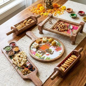 May include: A collection of wooden serving pieces including two rolling pin-shaped bowls filled with cookies, a small four-legged cot-style tray with nuts, a round board with an elephant illustration, and a long tray with dips and flatbread.