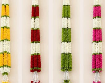 May include: Four colorful flower garlands hanging against a neutral background. The garlands are yellow, red, green, and pink, with white and green accents. Each garland has a tassel at the bottom and a loop at the top for hanging.