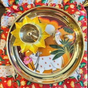 May include: A round brass plate featuring a central illustration of a white cow with orange spots, placed on a red fabric pouch with a repeating cow and lotus pattern. A yellow lotus-shaped brass diya and a small round lidded container are included.