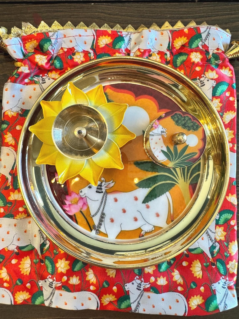 May include: A round brass plate featuring a central illustration of a white cow with orange spots, placed on a red fabric pouch with a repeating cow and lotus pattern. A yellow lotus-shaped brass diya and a small round lidded container are included.