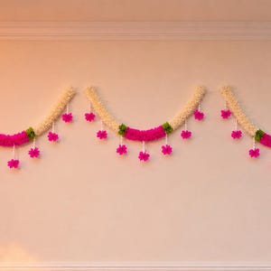 May include: Three decorative garlands hanging on a wall. Each garland has a cream-colored base with a vibrant pink center section and green accents. Pink flower-shaped ornaments are suspended from the garlands, adding a festive touch. The garlands are likely used for decoration.