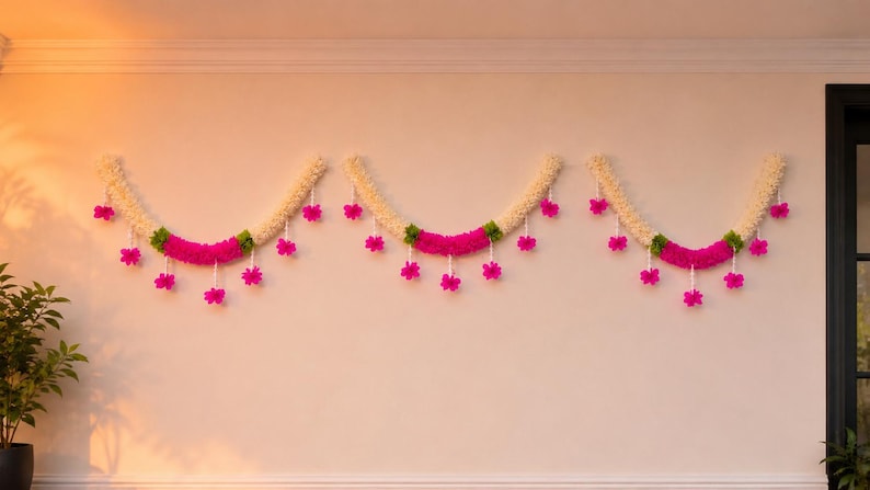 May include: Three decorative garlands hanging on a wall. Each garland has a cream-colored base with a vibrant pink center section and green accents. Pink flower-shaped ornaments are suspended from the garlands, adding a festive touch. The garlands are likely used for decoration.