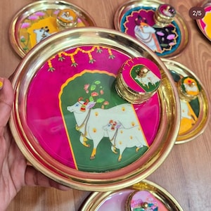 May include: A collection of decorative brass plates with lids, featuring vibrant hand-painted designs. The plates have a gold rim and a central image of a white cow and calf on a pink and green background. Other plates display similar artwork.