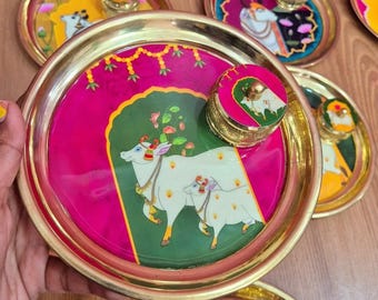 May include: A collection of decorative brass plates with lids, featuring vibrant hand-painted designs. The plates have a gold rim and a central image of a white cow and calf on a pink and green background. Other plates display similar artwork.