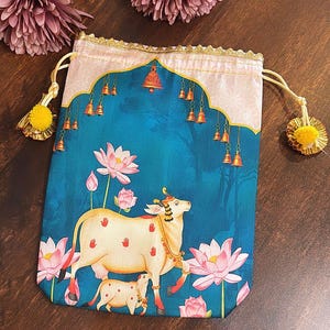 May include: A drawstring pouch with a teal background featuring a cow and calf illustration, pink lotus flowers, and hanging bells. The pouch has a gold trim and yellow tassels.