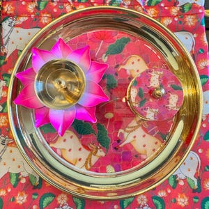 May include: A round gold-toned metal tray containing a pink and gold lotus-shaped candle holder and a small circular container with a pink cow-patterned lid, all resting on a matching pink fabric pouch featuring a repeating cow and lotus print.