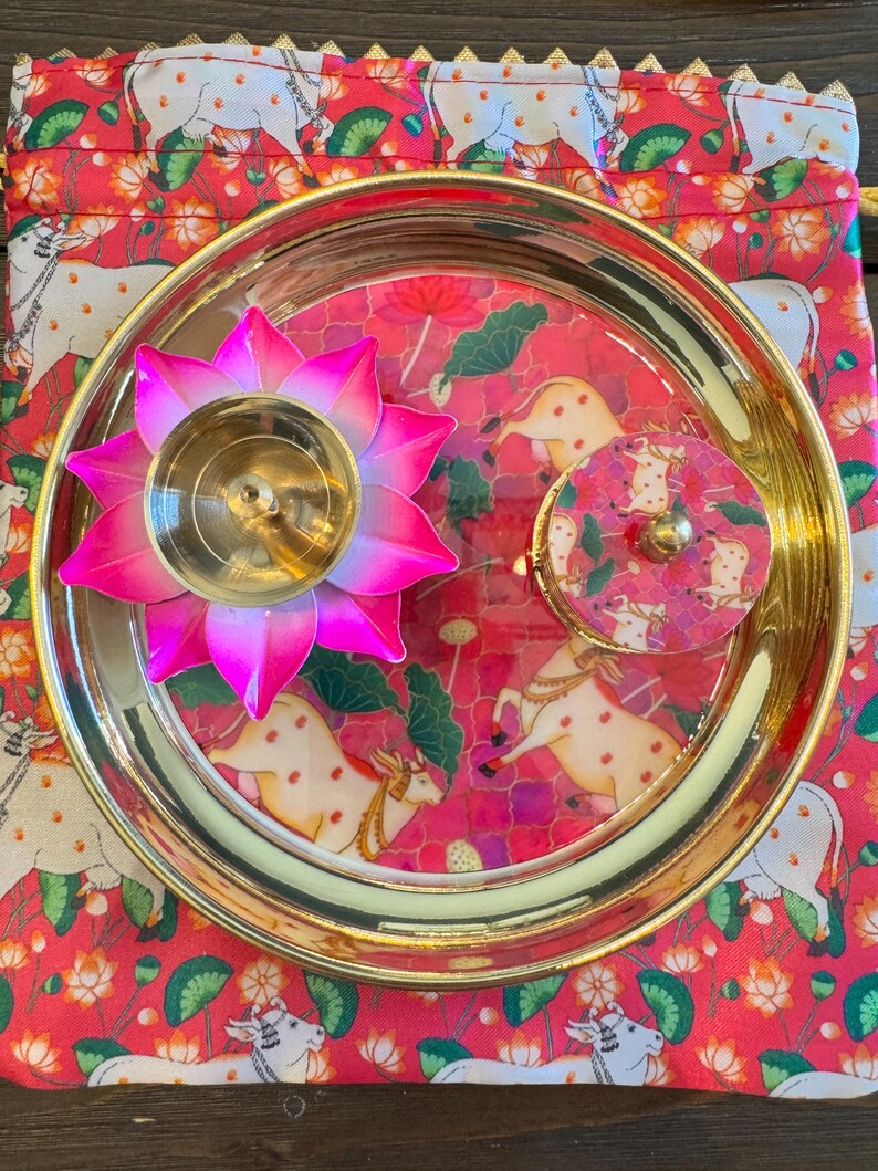 May include: A round gold-toned metal tray containing a pink and gold lotus-shaped candle holder and a small circular container with a pink cow-patterned lid, all resting on a matching pink fabric pouch featuring a repeating cow and lotus print.