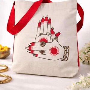 May include: A cream-colored canvas tote bag with bright red straps. The front features a graphic illustration of two hands positioned together, adorned with red nail polish, intricate red circular patterns, and detailed rings on the fingers.