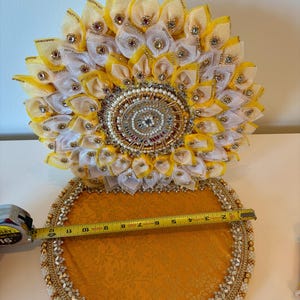 Puede incluir: Una bandeja circular decorativa con base de tela dorada y borde de cuentas, acompa&ntilde;ada de un adorno floral vertical con p&eacute;talos de tela amarillos y blancos, detalles dorados y pedrer&iacute;a. Una cinta m&eacute;trica muestra un di&aacute;metro de 30,5 cm.