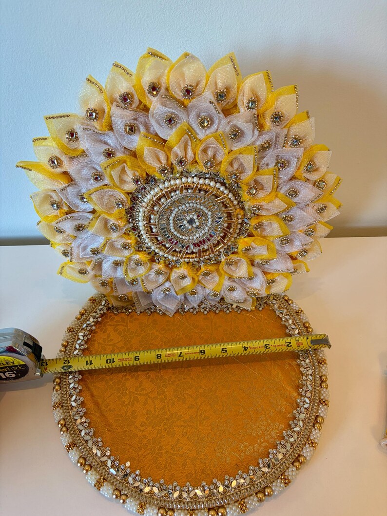 Puede incluir: Una bandeja circular decorativa con base de tela dorada y borde de cuentas, acompa&ntilde;ada de un adorno floral vertical con p&eacute;talos de tela amarillos y blancos, detalles dorados y pedrer&iacute;a. Una cinta m&eacute;trica muestra un di&aacute;metro de 30,5 cm.