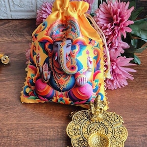 Op de afbeelding: Een gele zak met trekkoord met een levendige illustratie van de hindoeïstische godheid Ganesha. De tas is versierd met een kleurrijke afbeelding van Ganesha in een zittende houding. Een goudkleurige decoratieve lamp staat op de voorgrond.