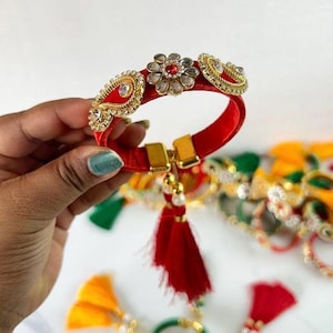 May include: A red fabric bracelet featuring a central flower-shaped embellishment with a red stone, flanked by two gold-toned paisley-shaped accents with clear crystals. The bracelet includes a gold-toned clasp and a dangling red tassel.