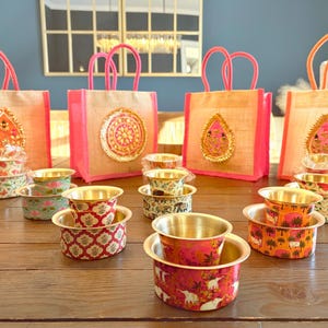 May include: Four jute gift bags with bright pink handles and decorative embroidered patches, arranged behind several small metal bowls. The bowls feature colorful floral, elephant, and geometric patterns with gold-toned interiors.