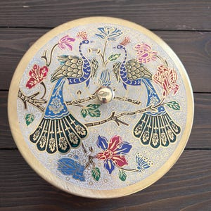 May include: Round, decorative metal container with a lid. The lid features a detailed design of two peacocks perched on a branch, surrounded by colorful floral patterns in blue, red, and gold. The container has a gold-colored rim and a small, round knob.