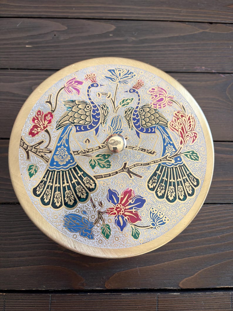 May include: Round, decorative metal container with a lid. The lid features a detailed design of two peacocks perched on a branch, surrounded by colorful floral patterns in blue, red, and gold. The container has a gold-colored rim and a small, round knob.