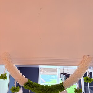 May include: A decorative garland featuring white and green floral elements. The garland is composed of white, fluffy sections and a central band of vibrant green, with hanging green flower clusters. It is suspended from a white ceiling.