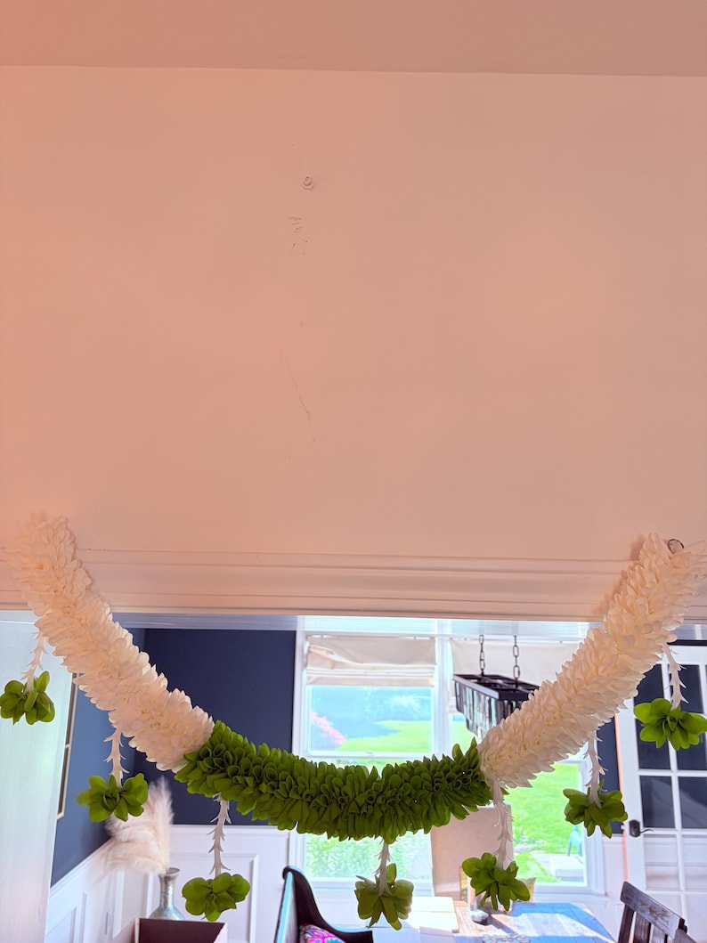 May include: A decorative garland featuring white and green floral elements. The garland is composed of white, fluffy sections and a central band of vibrant green, with hanging green flower clusters. It is suspended from a white ceiling.