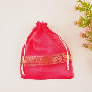 May include: A bright pink sheer organza drawstring pouch featuring a horizontal band of metallic gold woven floral embroidery across the center. The bag is cinched at the top with two thin white fabric cords that pull to close the opening.