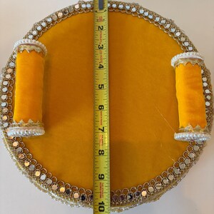 K&ouml;nnte beinhalten: Ein rundes gelbes Tablett mit zwei zylindrischen gelben Griffen, verziert mit einer Bord&uuml;re aus kleinen Spiegeln und wei&szlig;en Perlen. Ein gelbes Stanley-Ma&szlig;band in der Mitte zeigt, dass das Tablett einen Durchmesser von 25,4 cm hat.