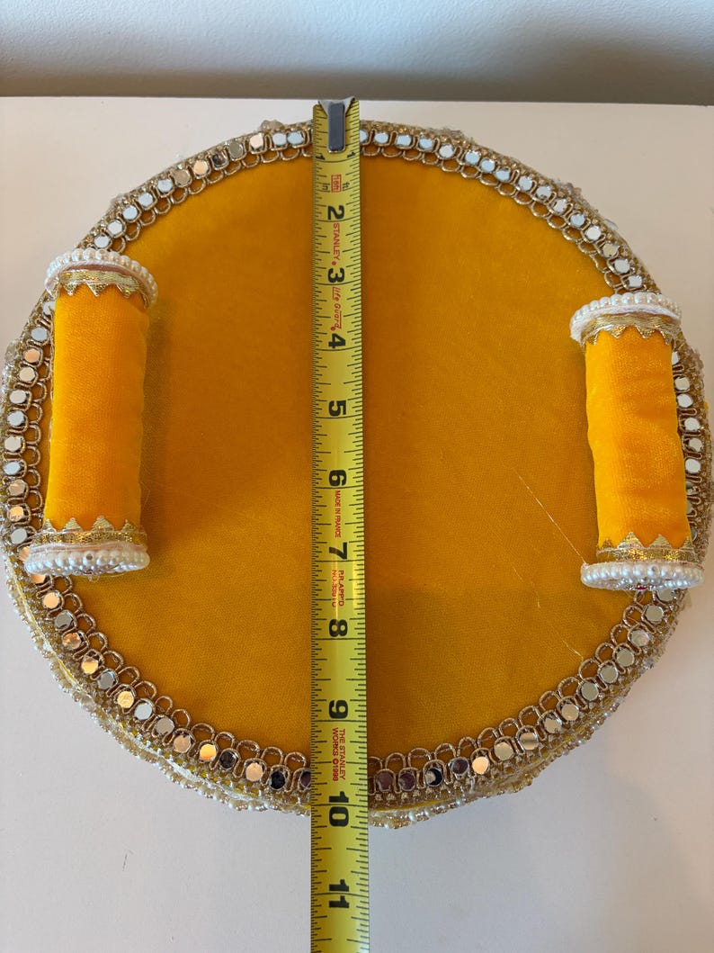 K&ouml;nnte beinhalten: Ein rundes gelbes Tablett mit zwei zylindrischen gelben Griffen, verziert mit einer Bord&uuml;re aus kleinen Spiegeln und wei&szlig;en Perlen. Ein gelbes Stanley-Ma&szlig;band in der Mitte zeigt, dass das Tablett einen Durchmesser von 25,4 cm hat.