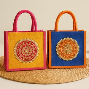 May include: Two square tote bags with sturdy handles. The left bag is yellow with pink trim and a red circular floral embroidery. The right bag is blue with orange trim and an orange circular floral embroidery, both featuring textured fabric.