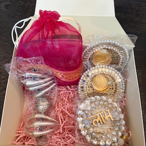 May include: A gift box containing a bright pink organza drawstring pouch, a silver-toned metal hanging ornament with curved elements, and three circular decorative candle holders adorned with clear crystals and the Hindi text "लाभ".