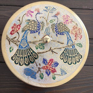 May include: A round, gold-colored decorative box lid with a detailed peacock design. The lid features two peacocks perched on a branch, surrounded by colorful floral patterns in blue, red, and pink. The lid has a gold knob.