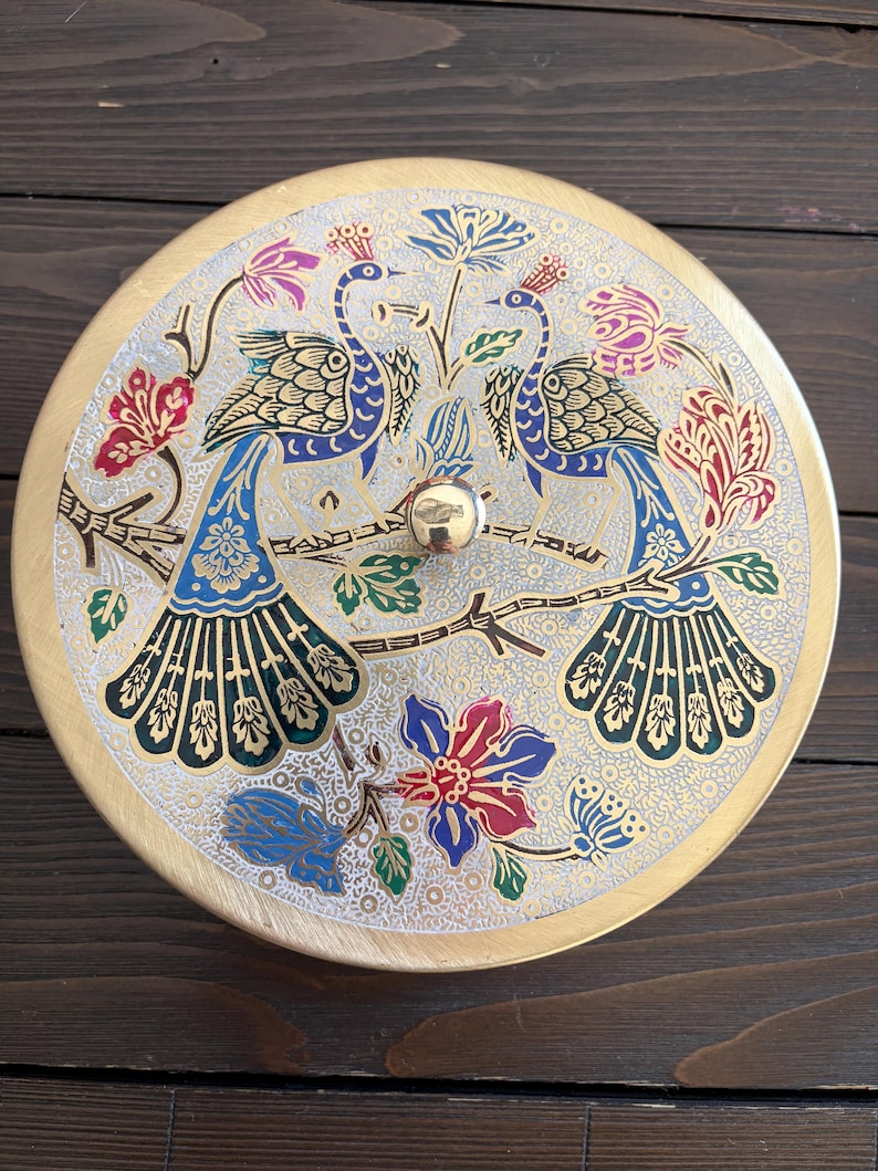 May include: A round, gold-colored decorative box lid with a detailed peacock design. The lid features two peacocks perched on a branch, surrounded by colorful floral patterns in blue, red, and pink. The lid has a gold knob.