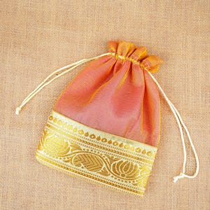 May include: A drawstring pouch made of iridescent coral-orange fabric featuring a wide, horizontal band of metallic gold woven trim at the bottom. The trim displays a detailed floral and paisley pattern. Two thin white cords cinch the top closed.