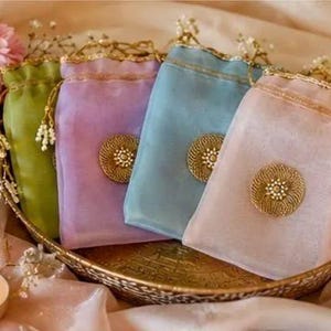 May include: Four rectangular drawstring pouches in olive green, lavender, light blue, and pale pink. Each sheer fabric bag features a gold circular medallion with a central floral design and gold drawstring cords with small white bead accents.