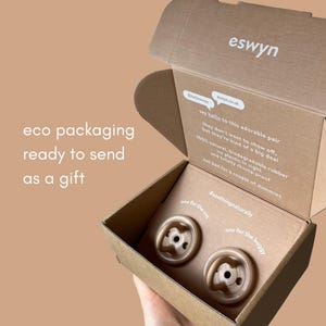 May include: A cardboard box with two beige baby pacifiers inside. The box is branded "eswyn" and states "eco packaging ready to send as a gift". The pacifiers are labelled "one for the cot" and "one for the buggy".