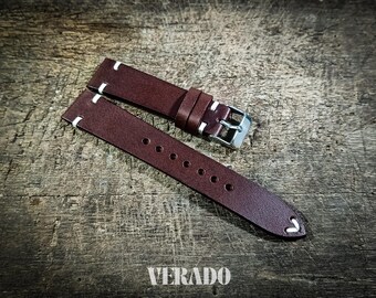 May include: A brown leather watch strap with white stitching and a silver buckle. The strap has multiple holes for adjustment and is displayed on a textured wooden surface. The word "VERADO" is at the bottom.
