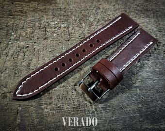 May include: A brown leather watch strap with white stitching and a silver buckle. The strap has multiple holes for adjustment and is displayed on a wooden surface. The word "VERADO" is printed at the bottom.